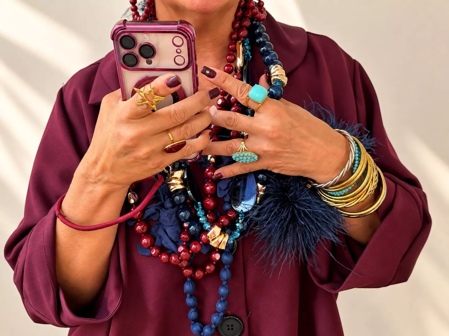 Femme rieuse en manteau pied-de-poule et lunettes de soleil, bracelets dorés au poignet, tenant un plateau débordant de colliers de perles blanches et dorées.