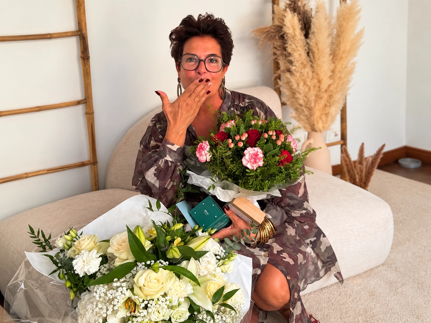 Femme assise sur un tabouret, veste en jean noir cloutée, pantalon fluide noir, lunettes carrées blanches, petit sac clouté et baskets à semelle épaisse.